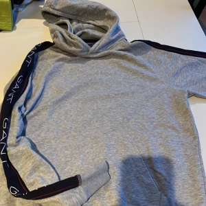 Grå hoodie från GANT - Säljer en snygg grå hoodie från GANT med logotyp längs ärmarna. Tröjan har en klassisk huva och en praktisk magficka. Perfekt för en avslappnad stil.
