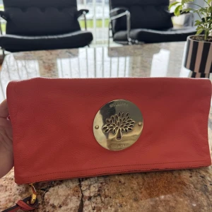 Röd kuvertväska från Mulberry - Snygg röd kuvertväska från Mulberry i skinn med en stor guldfärgad logotyp på framsidan. Väskan har en dragkedja inuti och en praktisk ficka med knappstängning. Perfekt för att bära med sig det viktigaste med stil.