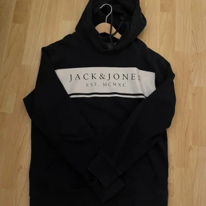 Svart/marinblå hoodie från Jack & Jones - Säljer en svart/marinblå hoodie från Jack & Jones med vit panel och text på bröstet. Den har en klassisk design med huva och långa ärmar. Perfekt för en avslappnad stil.