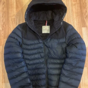 Svart dunjacka från Moncler - Säljer en snygg svart dunjacka från Moncler med huva och dragkedja. Jackan har en quiltad design och Moncler-logga på insidan. Perfekt för kyliga dagar.