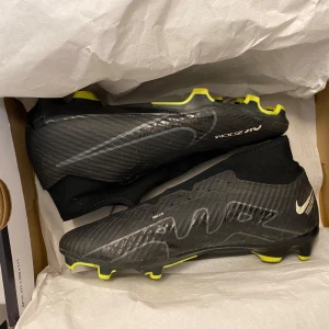 Svarta fotbollsskor från Nike - Snygga svarta Nike fotbollsskor med gula detaljer och en modern design. Skorna har en strömlinjeformad form med snörning och en diskret Nike-logga. Perfekta för fotbollsplanen med bra grepp och komfort.
