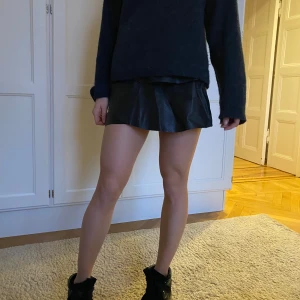 Svart kjol i skinnimaterial - Trendig svart kortkjol i skinnimitation med volangdetaljer och dragkedja bak. Perfekt för att ge din outfit en edgy touch. Passar bra till både sneakers och klackar.