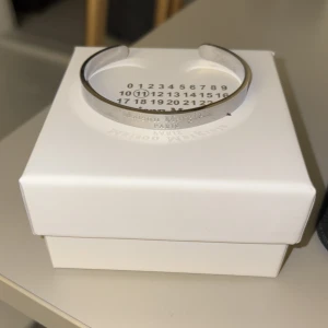 Silverfärgat armband från Maison Margiela - Elegant och stilrent silverfärgat armband från Maison Margiela. Armbandet har en minimalistisk design och kommer i en snygg förpackning med märkets ikoniska nummertryck. Perfekt för att ge en sofistikerad touch till din stil.