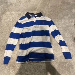Randig pikétröja från Polo Ralph Lauren - Snygg långärmad pikétröja från Polo Ralph Lauren i blått och vitt randigt mönster. Tröjan har en klassisk krage och knappar framtill. Perfekt för en stilren och avslappnad look.