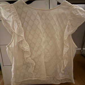 Vit blus med volangdetaljer - Säljer en söt vit blus med volangdetaljer längs ärmarna. Blusen har ett diskret mönster och är perfekt för en stilren look. Den är ärmlös och har en luftig passform. 