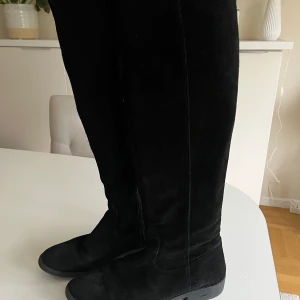 Svarta mockastövlar - Snygga svarta stövlar i mocka som går över knäna. Märke: Tamaris. Perfekta för en stilren look under höst och vinter. De har en klassisk design låg klack och en rund tå, vilket gör dem bekväma för dagligt bruk.