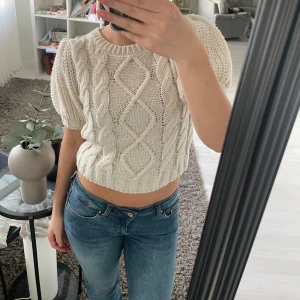 Vit stickad tröja med puffärmar - Säljer en söt vit stickad tröja med puffärmar och ett snyggt flätmönster framtill. Tröjan är croppad och har en rund halsringning. Perfekt för en stilren och mysig look. Passar bra till jeans eller kjol!