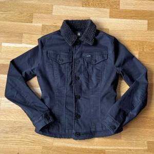 Dam G-Star Raw Svart Sherpa Trucker/Jeansjacka - Dam G-Star Raw Sherpa Trucker/Jeansjacka i färgen svart med sherpa foder och krage även med tryckknappar.  Storlek: XS, Armhåla till Armhåla: 46 cm, Längd: 55 cm, Axel till Armöppning: 61 cm. 
