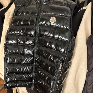 Svart dunväst från Moncler - Snygg svart dunväst från Moncler med glansig finish och dragkedja framtill. Perfekt för att hålla värmen med stil. Västen har en klassisk design med Moncler-logga på bröstet.