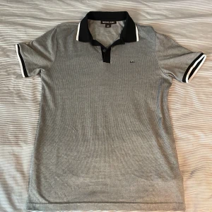 Grå pikétröja från Michael Kors - Snygg grå pikétröja från Michael Kors med svart krage och vita detaljer. Tröjan har korta ärmar och en liten broderad logotyp på bröstet. Perfekt för en stilren look.