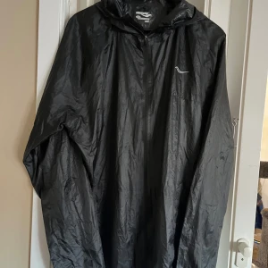 Svart vindjacka från Saucony - Säljer en svart vindjacka från Saucony i storlek XL. Jackan är tillverkad i 100% polyester och har en dragkedja framtill. Perfekt för blåsiga dagar med sin lätta och smidiga design.