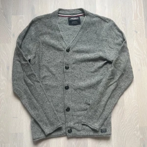 Grå kofta från Produkt - Snygg grå kofta från Produkt med knappar framtill. Den har en v-ringad design och långa ärmar, perfekt för kyligare dagar. Enkel och stilren, passar till många olika outfits.