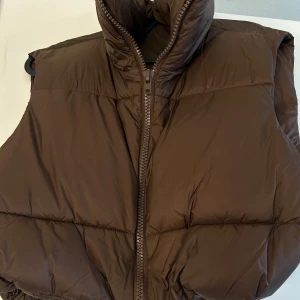 Brun dunväst med dragkedja - Säljer en brun dunväst med dragkedja. Västen har en hög krage och är perfekt för kyligare dagar. 