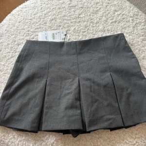 Grå kortkjol från Zara - Snygg grå kortkjol från Zara med veckad design. Perfekt för en stilren look. Kjolen är i en neutral grå färg som passar till det mesta. Märkeslappen sitter kvar vilket ger en känsla av nyhet.