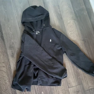 Svart hoodie från Polo Ralph Lauren - Säljer en klassisk svart hoodie från Polo Ralph Lauren med dragkedja och det ikoniska logotypbroderiet på bröstet. Tröjan har långa ärmar och en praktisk huva. Perfekt för en avslappnad stil. Laidback 