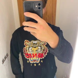 Svart sweatshirt från Kenzo - Säljer en svart sweatshirt från Kenzo med ett stort broderat tigerhuvud och texten 'Kenzo Paris' i färgglada toner på framsidan. Tröjan har långa ärmar och en rund halsringning. Perfekt för en stilren och trendig look.