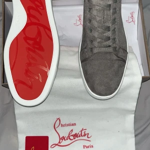 Grå sneakers från Christian Louboutin - Snygga grå sneakers från Christian Louboutin med den ikoniska röda sulan. Skorna har snörning och en stilren design som passar perfekt för en trendig look.