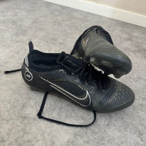 Svarta fotbollsskor från Nike - Snygga svarta fotbollsskor från Nike med vita och guldiga detaljer. Skorna har snörning och en modern design med fokus på hastighet och kontroll. Perfekta för fotbollsplanen med en stilren look.