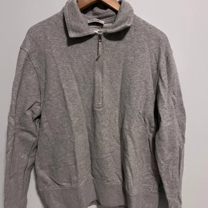 Grå tröja med dragkedja från COS - Säljer en s grå tröja från COS med halv dragkedja och hög krage. Perfekt för en avslappnad look. Tröjan har långa ärmar och är tillverkad i ett mjukt material.