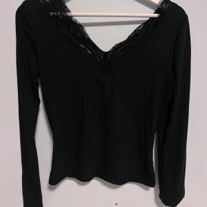 Svart ribbad topp med spetsdetaljer - Snygg svart ribbad topp med spetsdetaljer vid halsringningen. Toppen har långa ärmar och en v-ringad design som ger en elegant touch. Perfekt för att klä upp en outfit eller bära som den är.