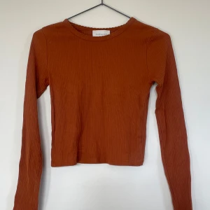 Orange ribbad långärmad topp -  Orange/brun ribbad topp med långa ärmar. Perfekt för höst och vår, den är både stilren och bekväm. Toppen är croppad och passar perfekt till ett par höga jeans eller kjol. Ett måste i garderoben för en trendig look! 🍂