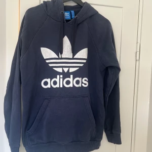 Mörkblå hoodie från Adidas - Säljer en snygg och bekväm mörkblå hoodie från Adidas. Den har en stor vit logga på framsidan och en klassisk känguruficka. Perfekt för både vardag och träning. Huvan har dragsko för justerbar passform. Passar perfekt till höst och vinter!