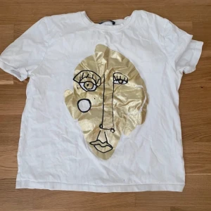 Vit t-shirt med guldtryck - Säljer en cool vit t-shirt med ett stort guldtryck av ett abstrakt ansikte på framsidan. T-shirten är kortärmad och perfekt för en avslappnad stil. Passar bra till både jeans och kjol för en trendig look! Små sminkfläckar vid kragen men de går bort i tvätten