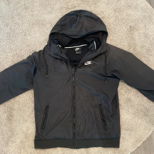 Svart vindjacka från Nike - Säljer en svart vindjacka från Nike i storlek S. Jackan har en dragkedja framtill och en justerbar huva. Den är perfekt för höst och vår, med långa ärmar och ribbade muddar vid ärmslut och nederkant. Den är i nyskick och använd vårterminen bara. 