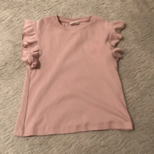 Rosa topp med volangärmar från Zara - Säljer en superfin rosa topp från Zara i storlek 130. Toppen har söta volangdetaljer vid ärmarna och är gjord i ett mjukt och bekvämt material. 
