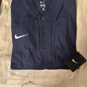 Mörkblå pikétröja från Nike - Säljer en snygg mörkblå pikétröja från Nike. Den har en klassisk krage med knappar och det ikoniska Nike-loggan broderad på bröstet. Perfekt för både vardag och träning! Storlek L passar M