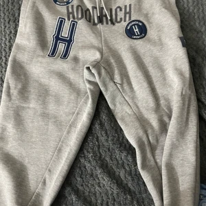 Hoodrich  - Säljer ett par grå mjukisbyxor från Hoodrich i storlek L. Byxorna har en avslappnad passform med snörning i midjan för justerbarhet. De är dekorerade med tryck och broderade detaljer i blått och vitt, vilket ger dem en sportig look. Perfekta för en c
