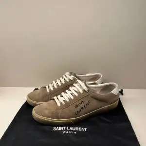 Säljer ett par snygga beige sneakers från Saint Laurent. De är i mocka med vita snören och har en vit detalj på hälen. Perfekta för en stilren look och passar till både vardag och fest. Skorna är i bra skick och kommer med originalpåse.