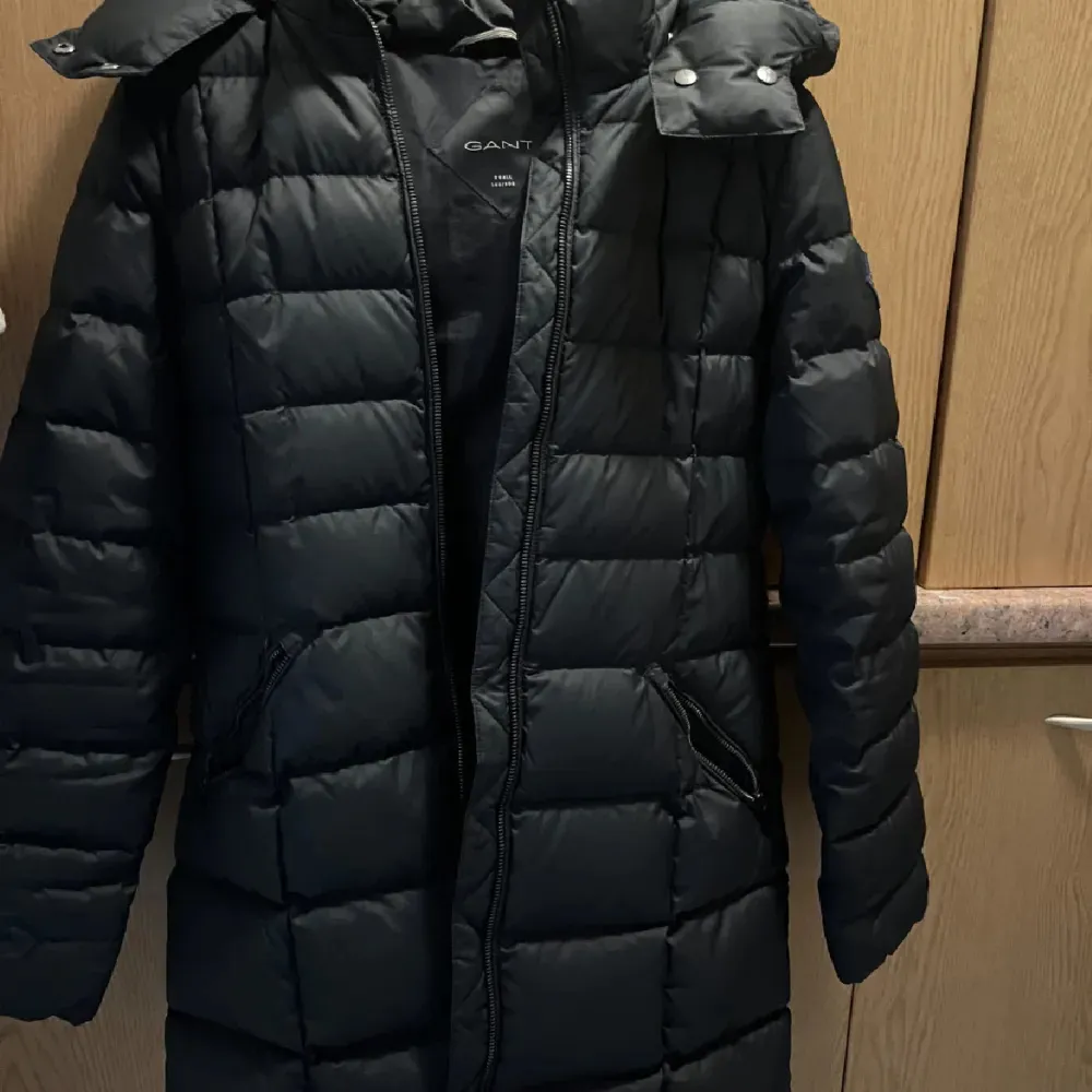 Säljer en svart dunjacka från GANT i storlek dam M. Jackan är lång och quiltad med dragkedja framtill. Perfekt för kalla vinterdagar med sin varma och mysiga design. Den har långa ärmar och en hög krage för extra skydd mot kylan.. Takit.
