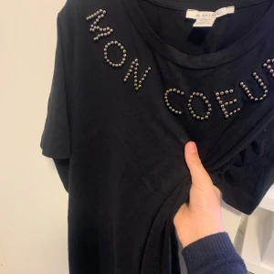 Svart t-shirt med pärldetaljer från Amisu - Säljer en svart t-shirt från Amisu i storlek XS. Den har coola pärldetaljer som bildar texten 'MON COEUR' på framsidan. Perfekt för en avslappnad men ändå stilren look. T-shirten är i bra skick och redo för nya äventyr!