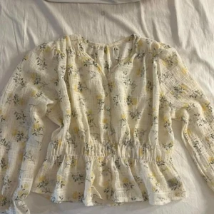 Blus - En söt blus med blommigt mönster och en elastisk midja. Blusen har långa ärmar med volanger vid ärmsluten och en knapprad framtill.säljer för 50