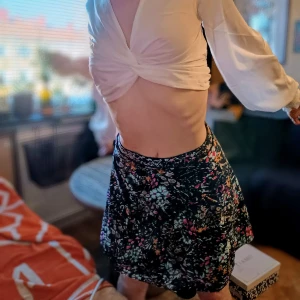 Blommig kortkjol - Säljer en superfin blommig kortkjol som är perfekt för sommaren! Kjolen har ett härligt mönster med olika färger som rosa, grön och vit på en svart bakgrund. Den är gjord i ett lätt och bekvämt material, vilket gör den idealisk för varma dagar. Passar perfekt till både vardag och fest!