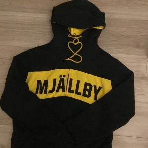 Mjällby hoodie  - Säljer en svart hoodie med gul detalj och Mjällby-tryck. Den har en dragkedja framtill och en mysig huva. Perfekt för kyliga dagar eller när du vill visa ditt stöd för Mjällby! 🖤💛