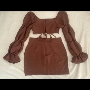 Brun tvådelad set med topp och kjol - Säljer ett snyggt brunt set med en croppad topp och matchande kjol. Toppen har långa puffärmar och knytning fram, vilket ger en trendig look. Kjolen är tight och kort, perfekt för en utekväll eller fest. Materialet känns mjukt och bekvämt. Passar perfekt för höst och vår!