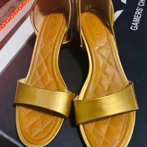 Guldiga sandaler med klack - Snygga guldiga sandaler från Ikatu med en bekväm klack. De har en stilren design med en bred rem över foten och en justerbar rem runt ankeln. Innersulan är quiltad för extra komfort. Perfekta för sommarens alla fester!