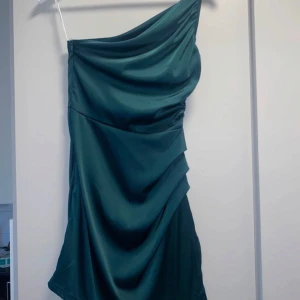 Grön oneshoulder klänning - Snygg grön oneshoulder klänning i ett elegant och flytande material. Perfekt för festliga tillfällen med sin stilrena design och draperade detaljer. Klänningen har en smickrande passform och är idealisk för både sommar och vår. Aldrig använd.