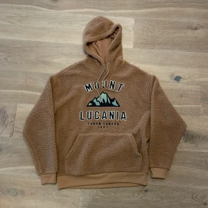 Brun fleece hoodie med tryck - Mysig brun fleece hoodie med texten 'Mount Lucania Yukon Canada 1897' tryckt på framsidan. Perfekt för kalla dagar med sin varma och fluffiga känsla. Den har en stor huva och ribbade muddar vid ärmslut och nederkant. Perfekt för höst och vinter!