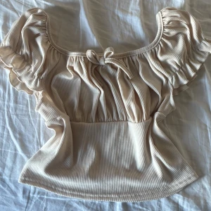Beige ribbad topp med volangärmar - Säljer en superfin beige ribbad topp med volangärmar och en söt knytdetalj framtill. Perfekt för sommardagar eller en mysig vårkväll. Toppen har en smickrande passform och är både bekväm och stilren.