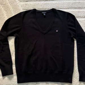 Svart ulltröja från GANT - Säljer en stilren svart tröja från GANT i 100% extrafin ull. Den har en klassisk v-ringning och är perfekt för både höst och vinter. Tröjan är i mycket bra skick och har en liten GANT-logga på bröstet. Perfekt för en elegant och bekväm look!