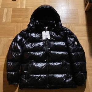 Svart dunjacka från Moncler - Säljer en snygg svart dunjacka från Moncler i nytt skick. Jackan är perfekt för vintern med sin pufferstil och långa ärmar. Den har en praktisk dragkedja och en mysig huva. Insidan har en cool serietidningsdesign. Perfekt för kalla dagar! 🖤