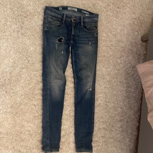 Blå jeansbyxor från Replay - Säljer ett par snygga blå jeansbyxor från Replay i modellen Anbass. De har en slim passform 28/32 och är i bra skick med slitningar som design. Perfekta för både vardag och fest! 