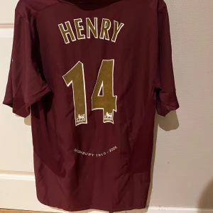 Vinröd Arsenal fotbollströja - Säljer en vinröd Arsenal fotbollströja från Nike med 'Henry' och nummer 14 på ryggen. Tröjan har korta ärmar och är i mycket bra skick. Den har ett guldigt O2-tryck på framsidan och ett Arsenal-märke på bröstet. Perfekt för samlare eller fans av Arsenal! ⚽
