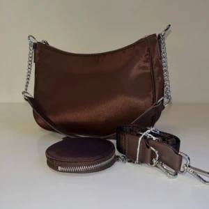 Brun axelväska med kedjedetaljer - Snygg brun axelväska i syntetmaterial med silverfärgade kedjedetaljer. Väskan har en avtagbar axelrem och en matchande rund plånbok med dragkedja. Perfekt för både vardag och fest! Helt oanvänd!