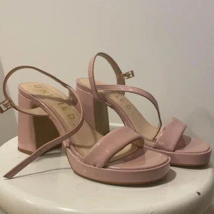 Rosa sandaler med klack - Snygga rosa sandaler från United med hög klack och justerbart spänne. Perfekta för sommarens fester eller en utekväll. De har en stilren design med bred rem över tårna och en smal rem runt ankeln.