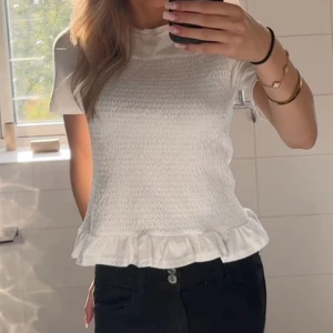 Vit topp med volangdetalj - Säljer en superfin vit topp med volangdetalj i nederkant. Toppen har korta ärmar och en smockad textur som är super snyggt❤️