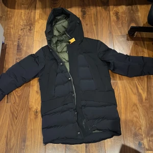 Svart dunjacka från Parajumpers - Säljer en svart dunjacka från Parajumpers i mycket bra skick. Jackan har en varm och vadderad insida i olivgrönt, perfekt för kalla vinterdagar. Den har en praktisk huva och stängs med dragkedja. Märkets logga finns på ärmen. Perfekt för dig som vill hålla stilen i vinter! ❄️